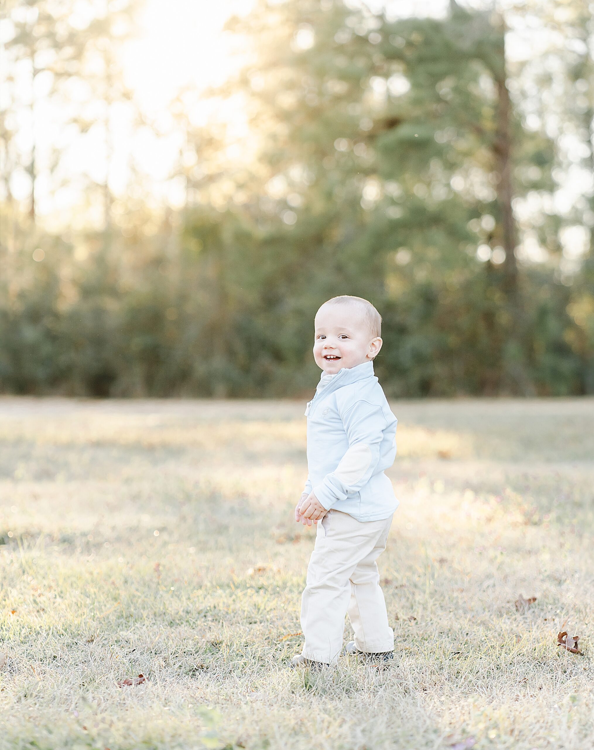 A one year old little boy stands and grins at the camera. Photo by Emily Lofton Photography.